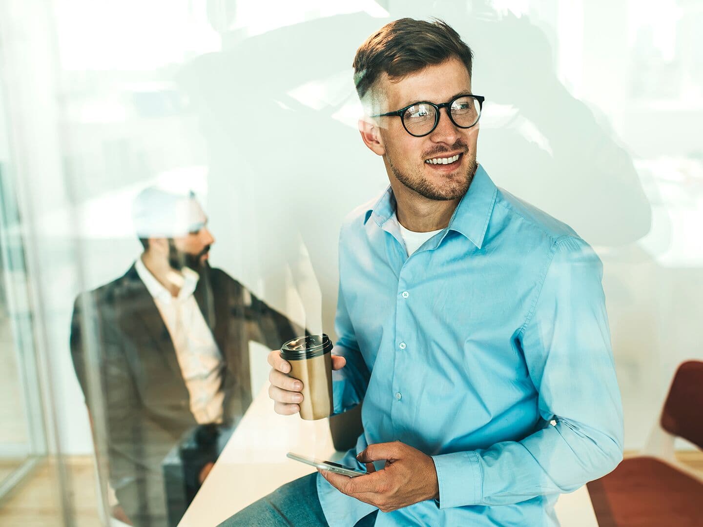 Man holding a cup of coffee with glasses inside an office with a phone on hand