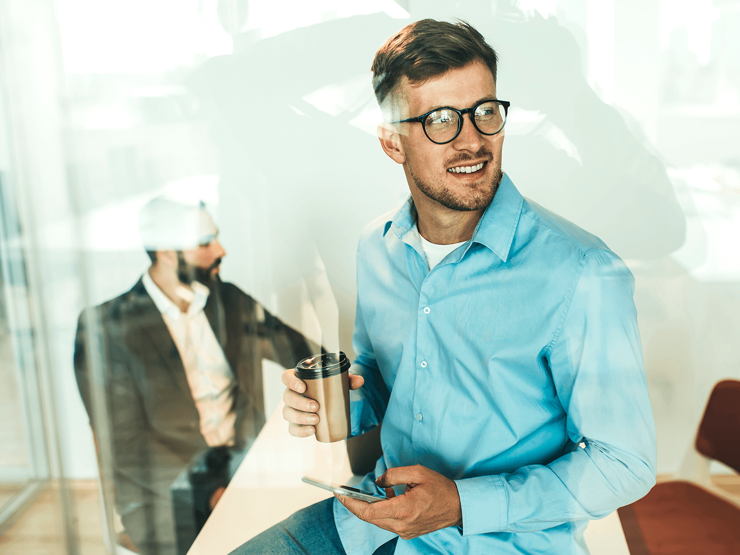 Man holding a cup of coffee with glasses inside an office with a phone on hand