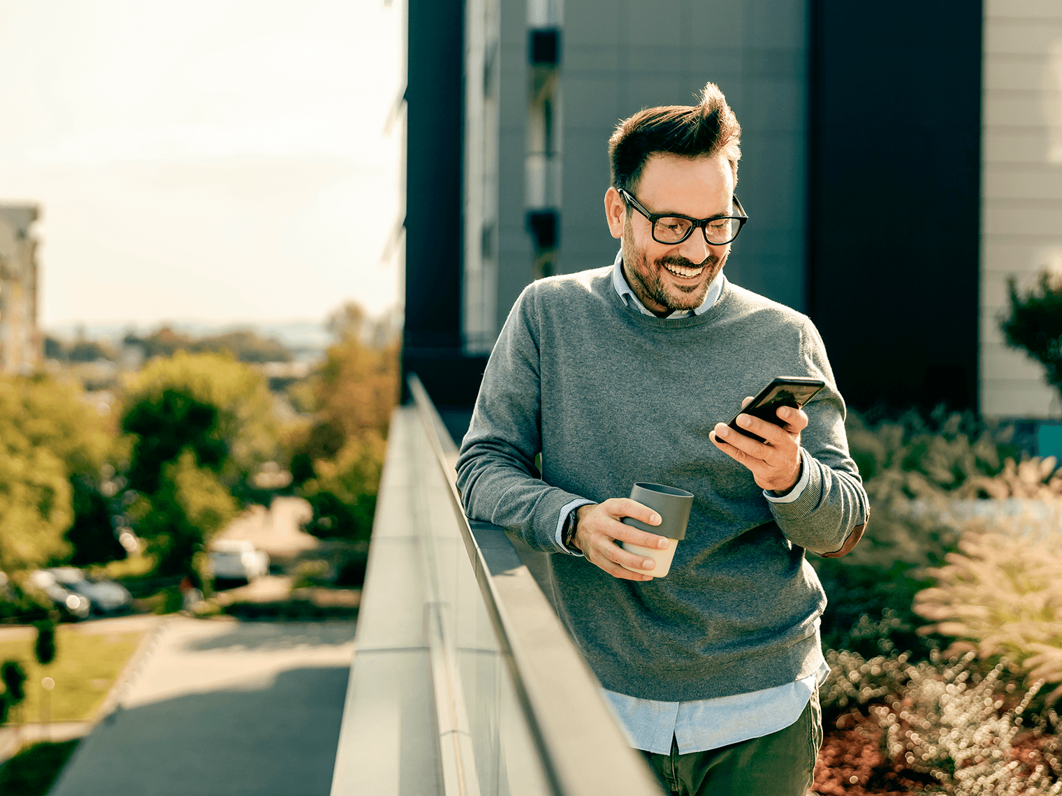 Man in glasses outdoors holding a mug and looking at his phone