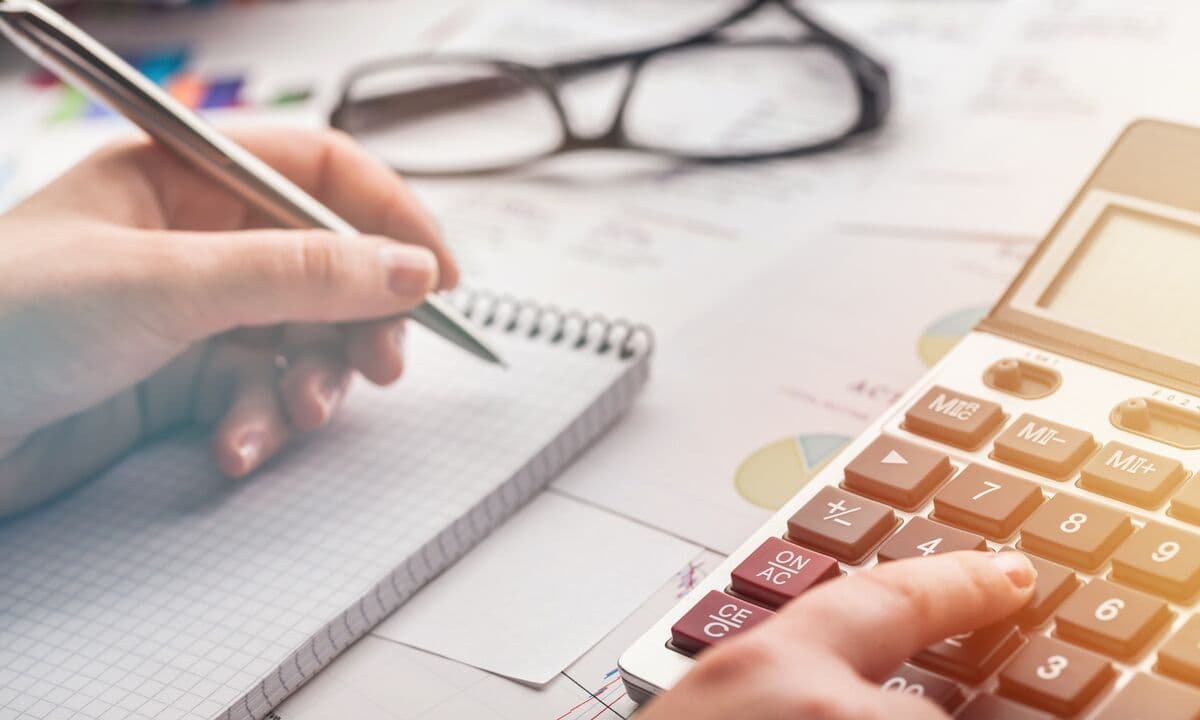 A person’s hand writing in a notebook next to a calculator on a desk.