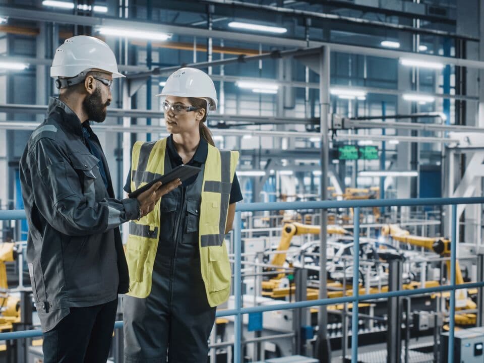 Two engineers wearing safety helmets standing in an industrial hall.