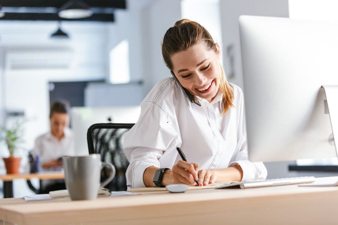 Cheerful young businesswoman sitting at her workplace at the office, talking on mobile phone, taking notes