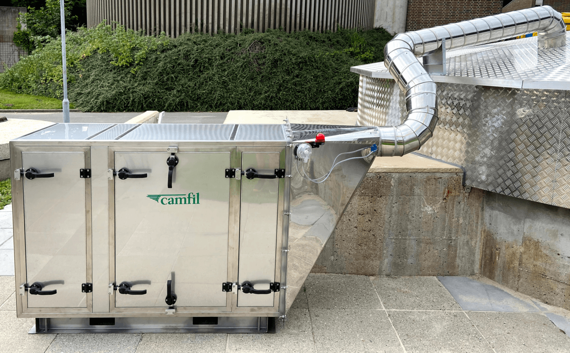 A stainless steel air filtration unit with multiple access doors and a connected ducting system, set on a concrete surface.