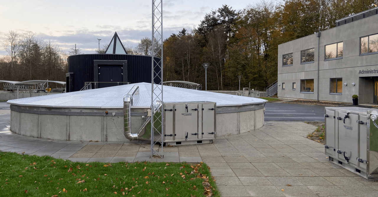 A modern facility with a circular structure, metal roof, and adjacent administrative building surrounded by trees and paved areas.